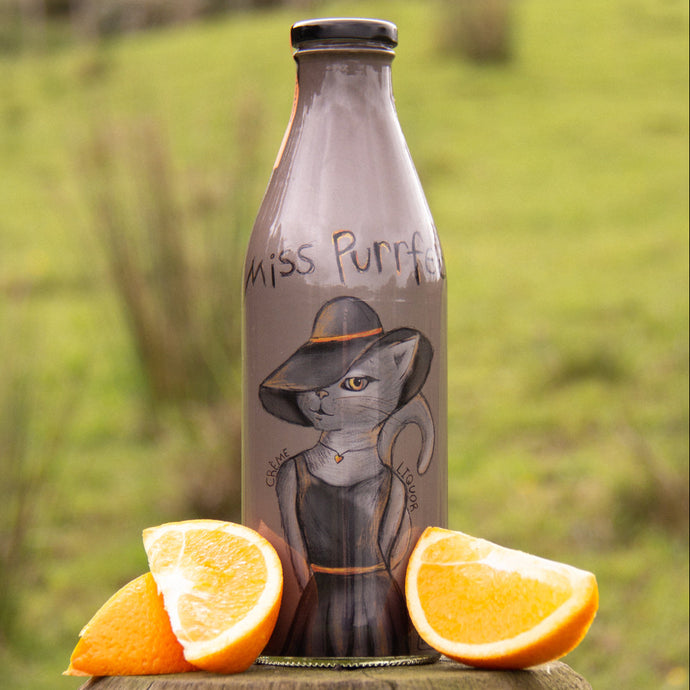Bottle with a cat illustration labeled 'Miss Purrfect' next to orange slices on a wooden surface with a grassy background