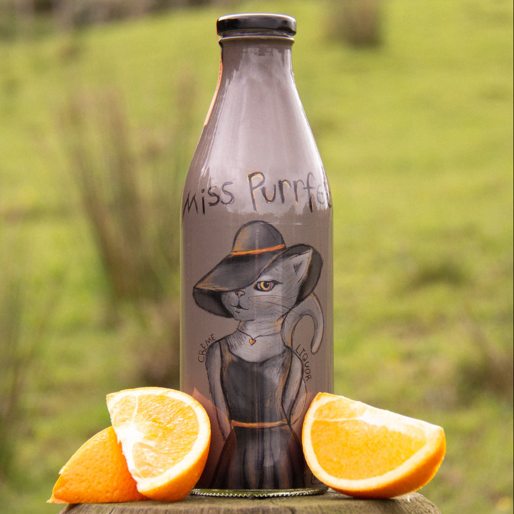 Bottle with a cat illustration labeled 'Miss Purrfect' next to orange slices on a wooden surface with a grassy background